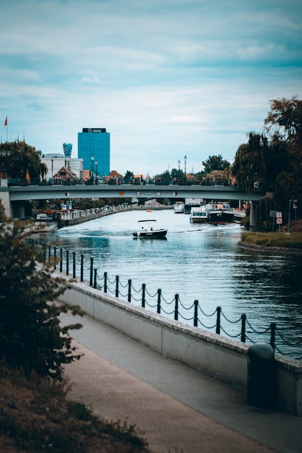 A view of a river in an urban setting shows a paved walkway with a black chain-link fence along the left side, bordered by green bushes and small trees. On the water, a small motorboat is passing beneath a bridge that spans the river, with several other boats visible in the background. The riverbanks are lined with residential and commercial buildings, with some trees and foliage nearby. In the background, a tall modern office building with a blue facade and a flagpole is visible, along with a few smaller structures. The sky overhead is partly cloudy with soft, diffused lighting, creating a calm and quiet atmosphere. This scene relates to the context of alternative waste handling methods, such as private rubbish collection services, with the river and urban environment emphasizing the importance of waste management in city areas. Rubbish Removal Putney operates within this setting, offering services that support effective rubbish disposal and collection for local residents and businesses in the SW15 area.