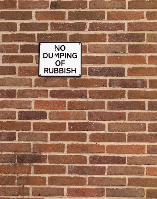 A close-up of a section of a red brick wall arranged in a horizontal running bond pattern with alternating rows of long and short bricks. The bricks have a rough texture with some variation in color, including shades of reddish-brown, dark brown, and light tan. Mounted on the wall is a rectangular white metal sign with black lettering that reads 'NO DUMPING OF RUBBISH.' The sign is centered within the image and positioned slightly above the middle, with visible mounting screws at each corner. The scene appears to be outdoors, with natural lighting casting soft shadows on the brick surface, indicating a sunny or partly cloudy day. The presence of the sign suggests a regulated or monitored area for waste management, aligning with the services provided by Rubbish Removal Putney, especially in the context of alternative rubbish collection and private waste handling methods.