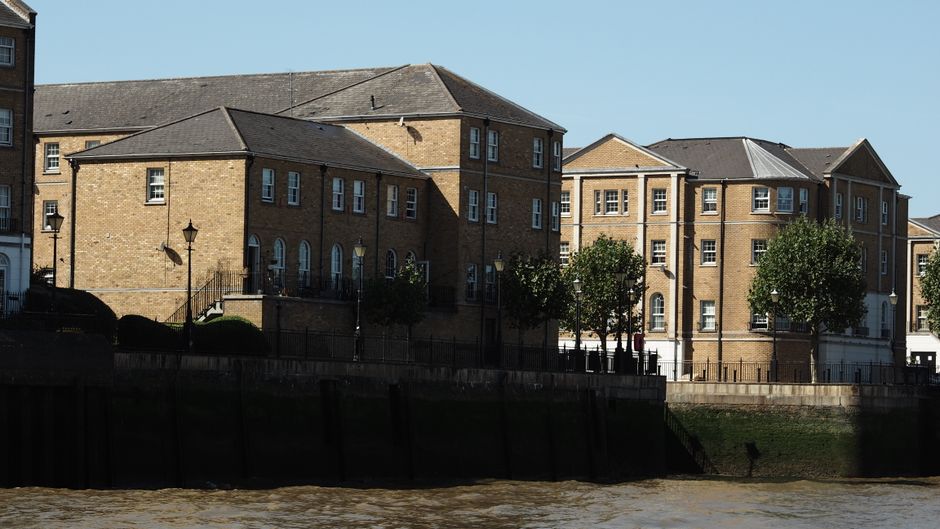 A row of multi-storey brick residential buildings situated along a riverside or canal edge, with a black metal fence separating them from the water. The buildings have light brown and beige brick facades, rectangular windows, and pitched roofs with dark shingles. Several small, leafy trees line the sidewalk in front of the buildings, casting shadows on the pavement. In the foreground, a dark wooden or concrete barrier runs parallel to the water, which has a rippled surface with a muddy brown tone. The scene is illuminated by natural daylight, with a clear blue sky overhead. The surroundings suggest a quiet urban riverside setting, where independent waste removal services, such as Rubbish Removal Putney, could facilitate rubbish collection from such residential properties engaging in private waste handling or on-site clearance, contributing to maintaining cleanliness along the riverside area.