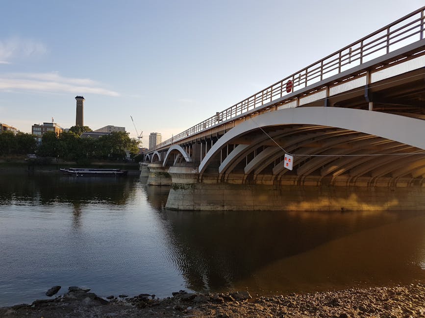 A wide view of a modern, white-painted bridge with curved arches spanning over a calm river during daylight hours. The bridge features a metal railing at the top and concrete supports extending into the water, with some visible rust and weathering. Beneath the bridge, the river reflects the structure and surrounding trees along the banks. To the left, a small boat is moored close to the riverbank, which is composed of rocks and soil. In the background, an urban skyline with various buildings, including a tall chimney or tower, and a construction crane, can be seen against a pale blue sky with minimal cloud cover. The scene is well-lit with natural light, emphasizing the clean lines of the bridge and the tranquil environment, which aligns with themes of infrastructure and potential private or independent waste management activities near riverside settings in urban areas. Rubbish removal services like those offered by Rubbish Removal Putney support maintaining such scenic and functional structures through effective refuse management.
