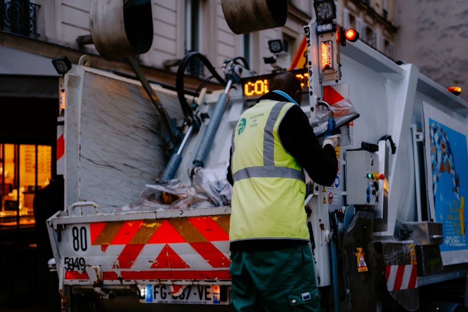 A waste collection operative wearing a black long-sleeved top, a high-visibility yellow vest with reflective stripes, and green trousers, is standing at the rear of a white rubbish bulk collection truck during dusk or evening. The truck is partially filled with large, mixed waste materials and has red and white reflective chevron markings on its rear. The worker appears to be operating controls on the side panel of the truck, which is equipped with various buttons, levers, and safety notices. The environment suggests an urban street setting, with buildings visible in the background, some with illuminated windows. The scene emphasizes the process of private waste handling or bulk rubbish collection, aligning with independent rubbish removal services. The lighting is subdued, with the truck’s indicator lights and surrounding ambient glow providing visibility, and the overall atmosphere conveys professional waste management activity consistent with rubbish removal operations in Putney.